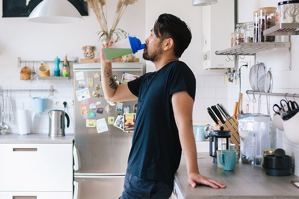 A man standing in kitchen and drinking green super drink from bottle.